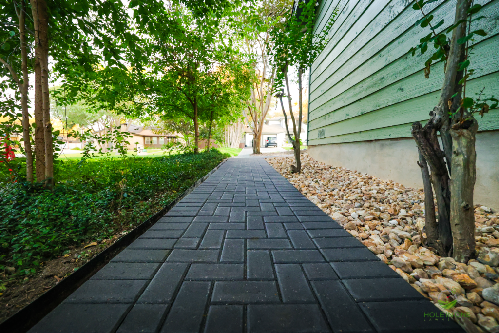 Long charcoal brick paver side-yard walkway between a hedge and green siding