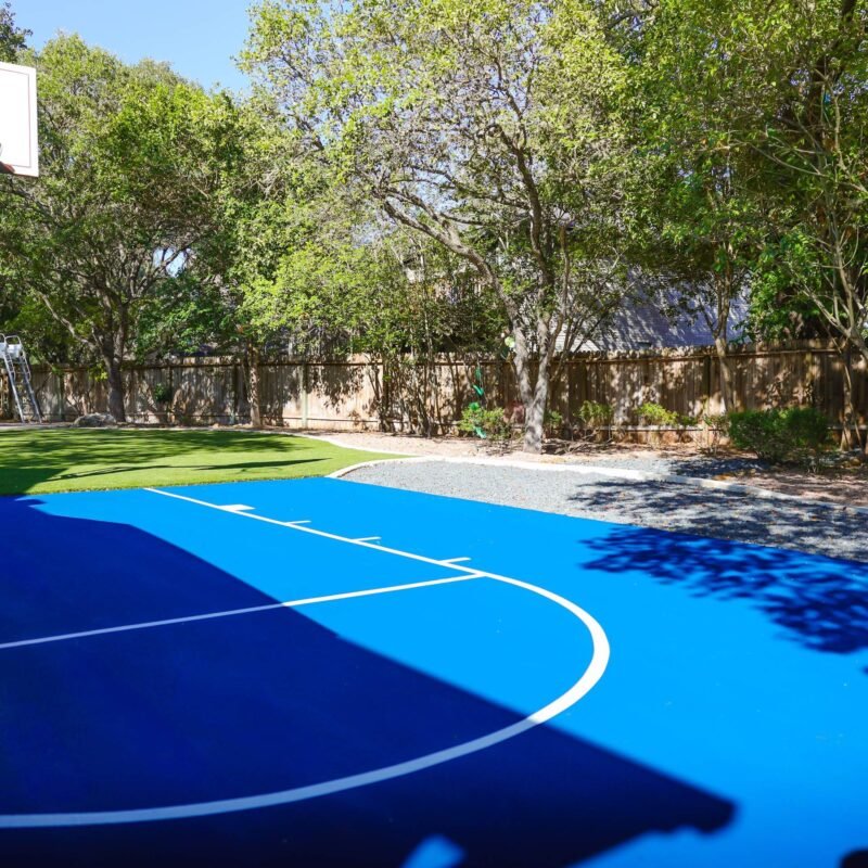 Blue acrylic backyard sport court with white game lines, basketball goal and artificial turf under mature oaks in San Antonio