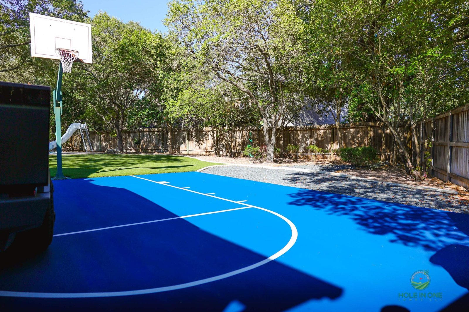 Blue acrylic backyard sport court with white game lines, basketball goal and artificial turf under mature oaks in San Antonio