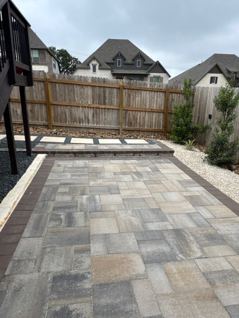 Multi-tone paver patio with dark soldier-course border beside a cedar privacy fence in a Boerne, TX yard