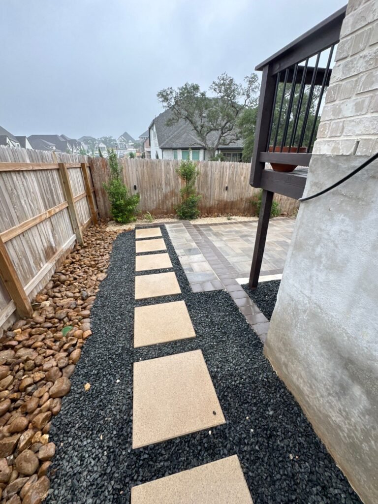 Side-yard path of square concrete stepping pads in black gravel with river rock border in Boerne, TX