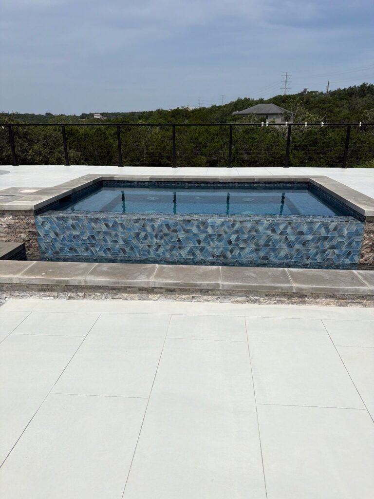 Mosaic-tiled spa with stone coping on a light porcelain paver pool deck in a Bulverde, TX backyard