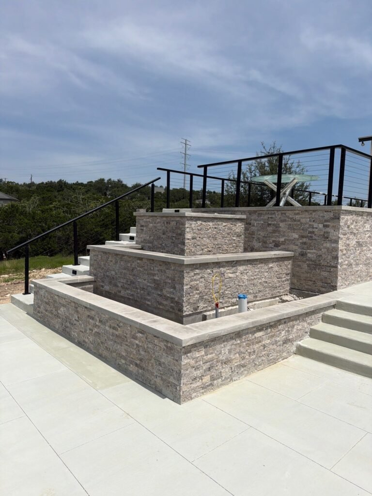 Tiered split-face stone veneer planter walls with steps and black metal railing in Bulverde, TX