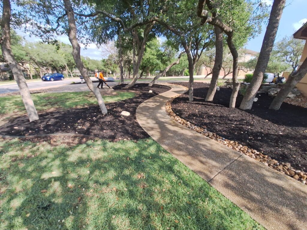 Curving front walkway lined with river rock and mulched tree beds under oaks in Garden Ridge, TX