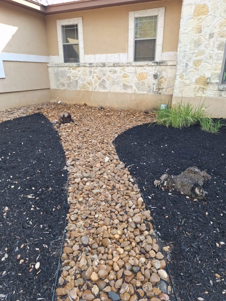 River rock dry-creek path between black mulch beds in front of a limestone home in Garden Ridge, TX