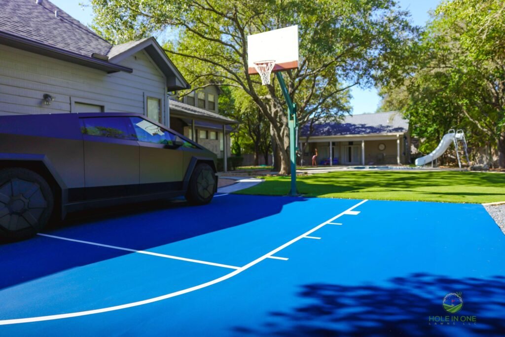 Finished blue sport court beside an artificial turf play lawn and slide, with gravel border and wood privacy fence