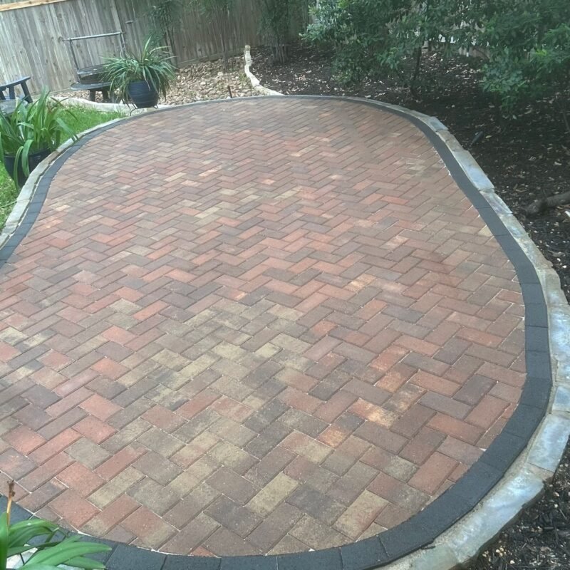 Oval herringbone clay-brick paver patio with charcoal border under mature trees in North San Antonio