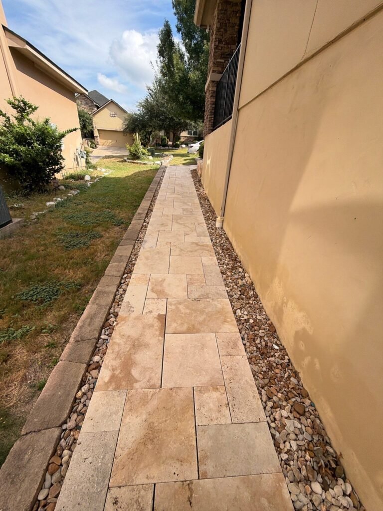 Side-yard travertine paver path bordered by river rock along a stucco wall in Stone Oak, San Antonio